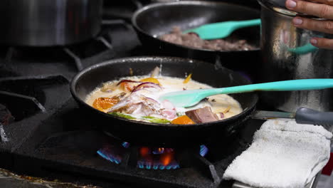 Chef-pours-ladle-of-coconut-milk-over-simmering-vegetable-curry-over-flame-in-commercial-restaurant-kitchen,-slow-motion-close-up-4K