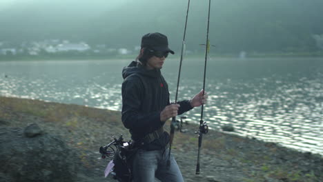 A-fisherman,-donning-sunglasses-and-a-cap,-prepares-to-cast-his-sleek,-modern-fishing-rod-into-the-tranquil-waters-of-Lake-Kawaguchiko-in-Yamanashi,-Japan-set-against-a-backdrop-of-quaint-white-houses
