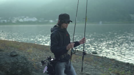 Un-Pescador,-Con-Gafas-De-Sol-Y-Una-Gorra,-Se-Prepara-Para-Lanzar-Su-Elegante-Y-Moderna-Caña-De-Pescar-En-Las-Tranquilas-Aguas-Del-Lago-Kawaguchiko-En-Yamanashi,-Japón,-Con-Un-Telón-De-Fondo-De-Pintorescas-Casas-Blancas.