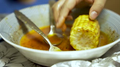 Corn-Cob-Being-Dipped-In-Spice-Sauce-Inside-Bowl