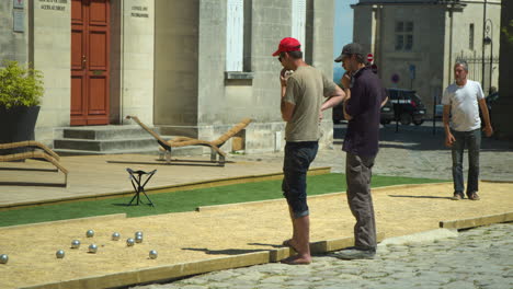 Adult-man-throwing-petanque-ball-on-gravel-field-while-his-two-friends-look