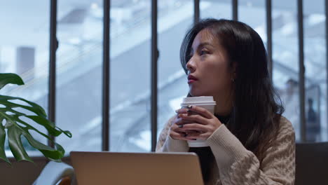 Nostalgic-young,-pretty-Asian-woman-sits-in-cafe-in-front-of-her-laptop,-holding-a-up-of-coffee,-thinking-about-life