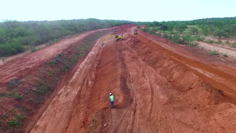 Excavator-at-work-in-a-red-soil-area-with-workers-and-trucks,-under-an-overcast-sky