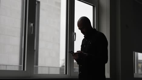 Silueta-De-Un-Hombre-Usando-Un-Teléfono-Inteligente-Junto-A-La-Ventana-En-Una-Habitación-Con-Poca-Luz,-Transmitiendo-Soledad