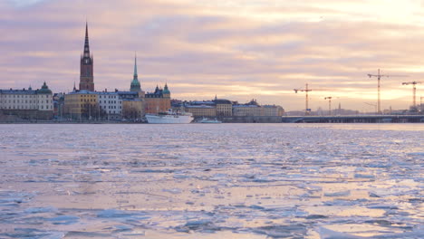 Gamla-Stan-Y-Slussbron-Contra-El-Cielo-Del-Atardecer,-Tiro-De-Seguimiento-Sobre-Agua-Congelada