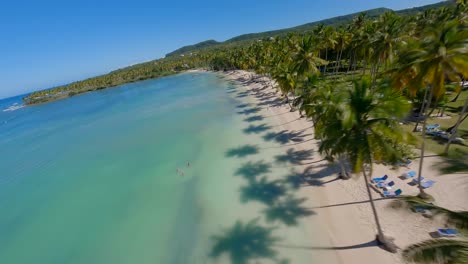 Drones-Se-Elevan-Sobre-La-Costa-Poco-Profunda-Mientras-Las-Palmeras-Proyectan-Hermosas-Sombras-Sobre-La-Arena-Dorada-Con-Turistas-Tomando-El-Sol,-Asserradero-Samana-República-Dominicana