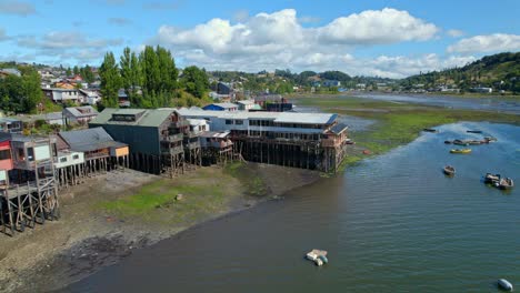 Chiloé's-stilt-houses-along-the-river,-colorful-boats,-sunny-day,-chilean-culture,-aerial-view