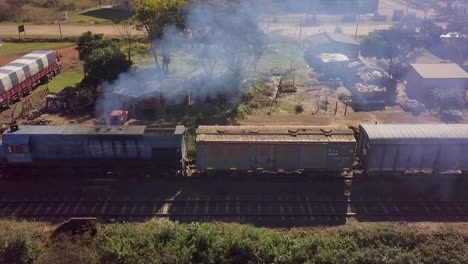 An-argentine-train-passing-through-a-rural-area,-smoke-rising,-houses-nearby,-sunny-day,-aerial-view