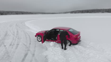 Man-using-shovel-to-dig-out-his-red-car-from-snowbank-at-ice-race-track