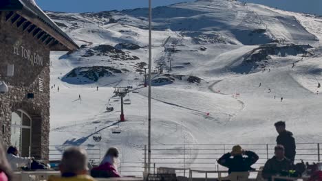 A-group-of-people-on-a-sunny-ski-restaurant-terrace-after-a-day-of-skiing,-watching-the-skiers-on-the-slopes