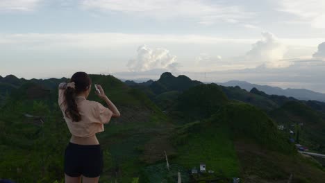 Süße-Frau-In-Bauchfreiem-Top-Und-Shorts-Ordnet-Ihr-Haar,-Während-Sie-Bei-Sonnenuntergang-Auf-Die-Atemberaubende-Aussicht-Auf-Die-Tropischen-Berge-Blickt