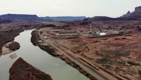 A-high-flying-drone-shot-of-an-industrial-mining-complex-and-a-railroad-track-running-along-the-Colorado-River,-cutting-through-the-unique-and-rugged-desert-landscape-near-Moab,-Utah