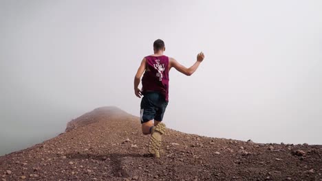 Low-angle-follow-cam-of-a-black-haired-man-in-shorts-and-a-tank-top-running-on-an-isolated-cloud-covered-rocky-mountain-ridge