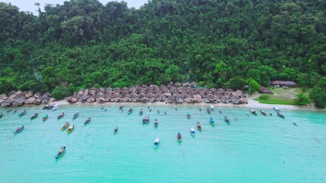 Aerial-Drone-Pull-Out-Shot-Over-Long-Tail-Boats-at-a-Wooden-Hut-Village-Along-a-Tropical-Beach-in-Thailand-with-Green-Hillside-in-the-Background
