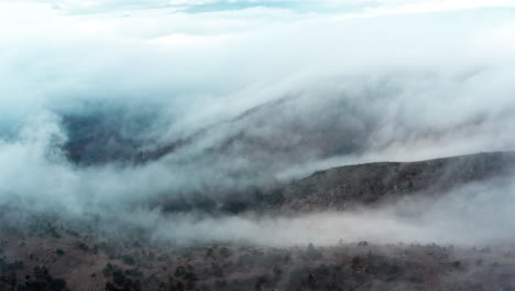 Misty-clouds-rolling-over-a-forested-hillside,-creating-a-serene,-mysterious-atmosphere,-aerial-view
