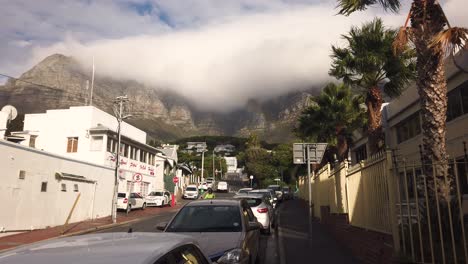 Wind-blows-through-palm-trees-in-Camp's-Bay,-South-Africa,-with-mist-over-Table-Mountain-visible-in-background