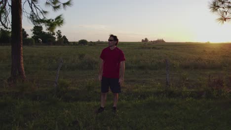 Young-man-looking-at-horizon-amidst-green-fields-and-trees-at-sunset