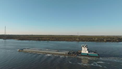 An-aerial-view-of-a-tugboat-pushing-a-barge-up-the-Houston-Ship-Channel-before-sunset-under-clear-blue-skies-in-Houston,-Texas