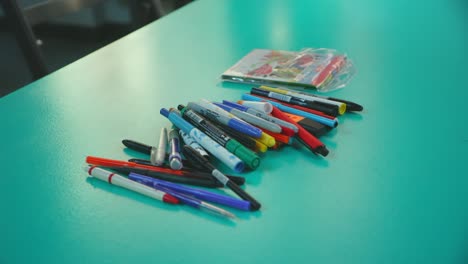 Bolígrafos,-Lápices-Y-Marcadores-De-Diversos-Colores-En-Un-Primer-Plano-De-Un-Estante-De-Enfoque-De-Lente-De-Mesa-Verde