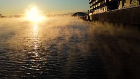 Aerial-Low-Flying-Over-Calm-Waters-Beside-Waterside-Apartments-With-Floating-Mist,-Tilt-Up-To-Reveal-Golden-Hour-Sunset-Near-Bjorvika,