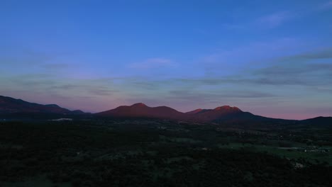 Rückzugsflug-In-Einem-Tal-Mit-Einem-Großen-Waldgebiet-Und-Ländlichen-Bauernhöfen-Bei-Sonnenuntergang,-Der-Die-Strahlen-Auf-Einige-Berge-Wirft-Und-Ihnen-Einen-Orangefarbenen-Ton-Und-Einen-Farbenfrohen-Himmel-Im-Winter-In-Avila,-Spanien-Verleiht