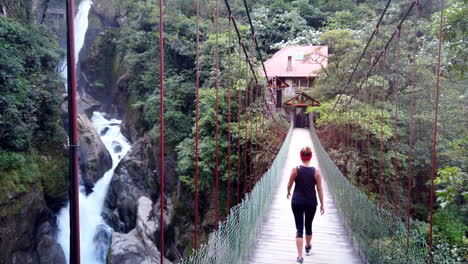 Mujer-Caminando-Hacia-Una-Cabaña-Sobre-Un-Puente-Colgante-En-Baños,-Ecuador,-Rodeada-De-Exuberante-Vegetación-Y-Un-Río-Caudaloso-Debajo