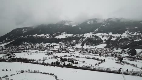 Aerial-gloomy-view-of-snowy-countryside-field-near-cloud-covered-mountain