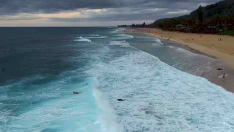 Aerial-view-of-waves-crashing-over-surfers-at-Ehukai-Beach-Park-shore,-Ohau