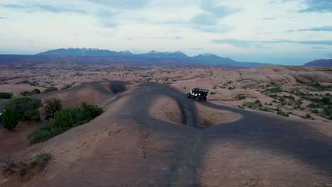 A-4K-tracking-drone-shot-of-a-lifted-jeep-off-roading-through-the-extreme-and-rocky-desert-landscape-near-Moab,-Utah,-with-the-snowy-rocky-mountains-towering-in-the-distance