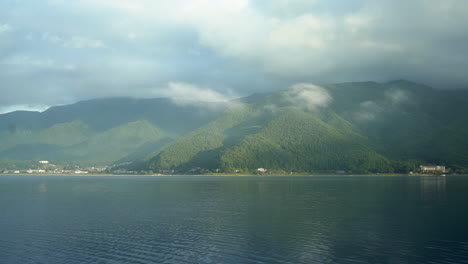 The-stunning-Lake-Kawaguchiko-in-Honshu,-Japan-showcases-lush-green-mountains-in-the-background-and-fluffy-clouds,-reflecting-buildings-on-its-tranquil-waters,-epitomizing-the-serenity-of-the-lake