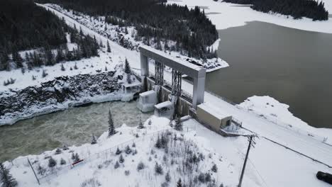 Long-Snow-Covered-Road-with-Aerial-Slow-Drone-Orbit-Rushing-Water-Notigi-Hydroelectric-Dam-in-the-Arctic