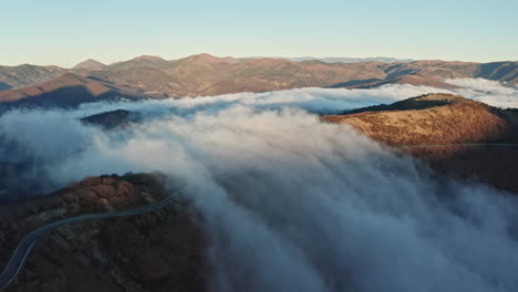 Nubes-Rodando-Sobre-El-Paisaje-Montañoso-Al-Amanecer,-Camino-Sinuoso-A-Través-De-Colinas,-Vista-Aérea