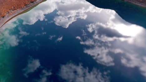 Lavodilec-lake's-turquoise-waters-with-clouds-reflecting,-mountainous-terrain,-aerial-view