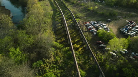 Aerial-shot-of-a-train-passing-by-a-junkyard-in-Fayetteville,-AR-during-the-day,-sunlight-touching-treetops