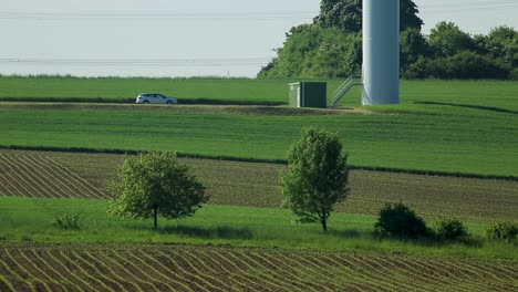 Car-parked-by-a-wind-turbine-in-sunny-rural-landscape,-green-fields,-serene-day,-distant-power-lines