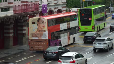 Vehicle-traffics-on-hill-street-downtown-Singapore,-carbon-footprint-public-transportation,-electric-double-decker-buses-and-commuting-cars-driving-on-the-road-on-a-raining-day-in-the-city-center