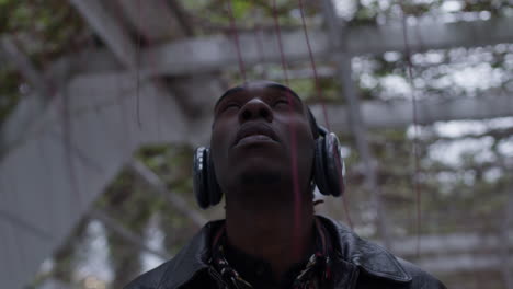 Aspired-young-black-man-with-fashionable-leather-jacket-listens-to-music-in-headphones-outdoors,-parallax-shot