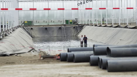 Dos-Hombres-Caminando-Dentro-Del-Estanque-De-Riego-Del-Invernadero,-Sitio-De-Construcción-De-Nuevas-Tuberías