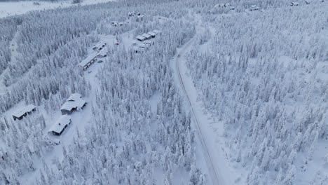 Vista-Aérea-Rodeando-Un-Coche-Ev-En-Medio-De-Un-Bosque-Nevado-Y-Cabañas-De-Laponia