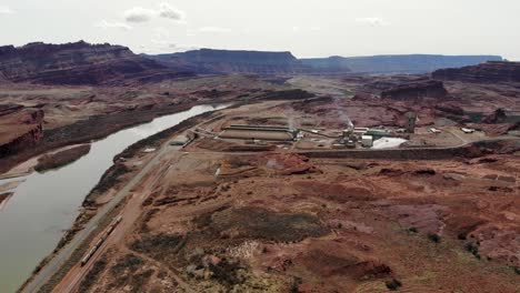 A-high-flying-drone-shot-of-an-industrial-mining-complex-and-a-railroad-track-running-along-the-Colorado-River,-cutting-through-the-unique-and-rugged-desert-landscape-near-Moab,-Utah