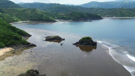 Drone-flyover-shot-of-stunning-low-tide-beach-and-large-rocks-with-crashing-ocean-waves-and-lush-island-hills-background