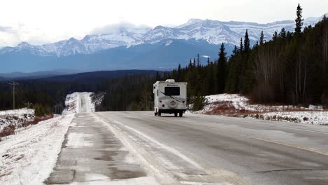 Camper-van-driving-down-a-snowy-winter-road-heading-towards-the-snow-covered-mountains