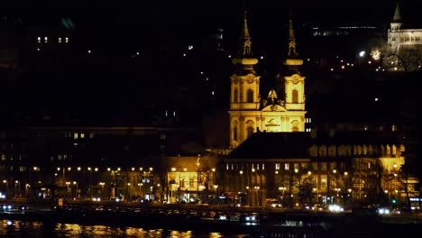 Vista-Del-Centro-De-La-Ciudad-De-Budapest-Con-La-Torre-De-La-Iglesia-Iluminada-Y-El-Río-Danubio-Por-La-Noche,-Arquitectura-Gótica,-Reflejos-De-Luz,-Toma-Distante