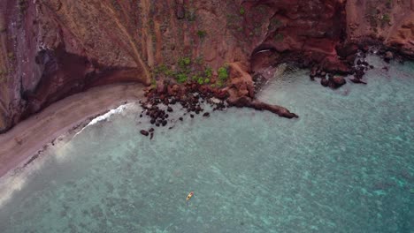 Cinematic-aerial-view-capturing-tourists-rowing-in-a-colorful-kayak-in-tropical-Hawaiian-exotic-destination-with-turquoise-calm-ocean-surface-and-red-dirt-cliffs