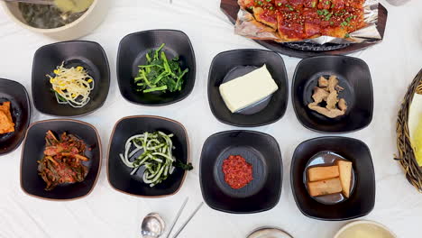 Top-down-view-of-a-traditional-Korean-meal-with-spicy-marinated-fish,-various-side-dishes-including-kimchi-and-greens,-tofu,-and-seaweed-soup,-all-neatly-presented-on-white-table---crane-up-reveal