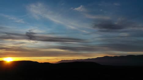 flight-in-a-sunset-in-a-valley-with-a-mountain-system-of-dark-almost-black-color-with-a-yellow,-blue-sky-and-with-gray-clouds-and-the-sun-setting-in-the-mountains-watching-it-with-a-twist-in-Spain