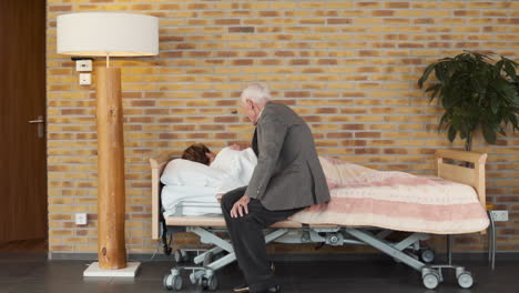 Elderly-man-sitting-down-on-bed-side-to-comfort-woman-sleeping,-nursing-home