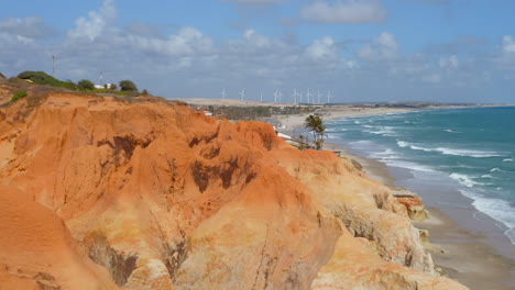 Aerial-view-of-the-sea,-waves,-cliffs,-a-small-village-and-a-wind-energy-at-background,-Morro-Branco,-Ceara,-Brazil