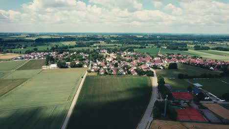 A-quaint-village-surrounded-by-lush-green-fields-under-a-blue-sky,-aerial-view