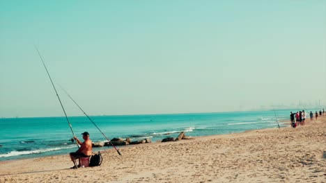 Static-Shot-of-Old-Shirtless-Man-Fishing-on-the-Beach,-People-in-Background